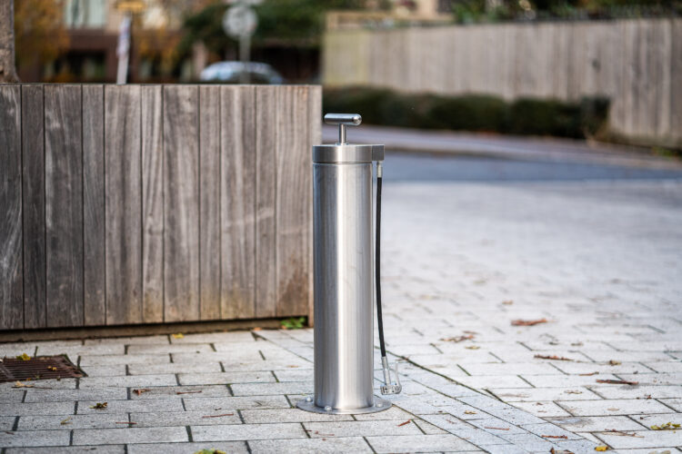 A cylindrical metal Public Bike Pump affixed to a paved footpath.
