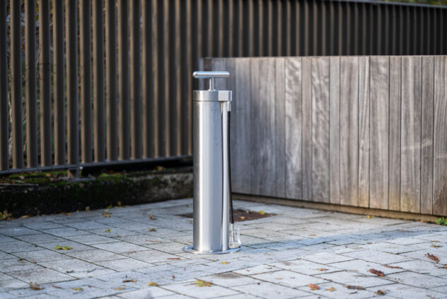 A tall, metal, cylindrical public bike pump affixed to the paved ground, surrounded by wood panelling.